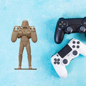 May include: A tan Stormtrooper-shaped controller holder stands next to a black and a white PlayStation controller. The controllers have buttons and joysticks. The background is a textured light blue surface.