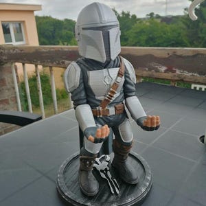 May include: A grey and silver 3D printed figurine of a Mandalorian character from the Star Wars franchise. The figurine is standing on a black circular base with a Mandalorian symbol.