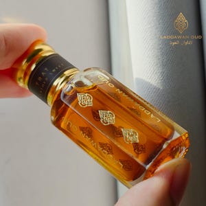Op de afbeelding: Een kleine, rechthoekige parfumfles gevuld met amberkleurige vloeistof. De fles heeft een gouden dop en is voorzien van de tekst "Laddawan Oud" in gouden letters. Het etiket bevat ook de tekst "TRAT Leather Sweet".