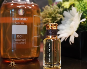 Może przedstawiać: Przezroczysty szklany flakon perfum ze złotą nakrętką i napisem "LADDAWAN OUD" jest prezentowany obok dużego szklanego zlewki. Flakon perfum ma ciemnobrązową etykietę ze złotymi literami. Tło obejmuje kompozycję kwiatową.