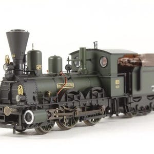 May include: A green and black model train locomotive with a brown wooden caboose. The locomotive has a large chimney and a number "18" on the side. The caboose has the text "Die Gruben Bahn" on the side.