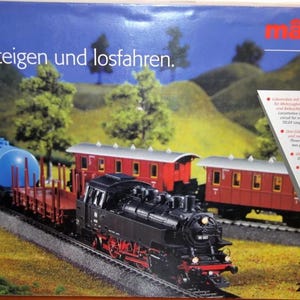 Puede incluir: Un set de tren modelo Märklin a escala HO con una locomotora de vapor negra que tira de tres vagones de pasajeros rojos y un vagón cisterna azul. La caja presenta el texto "Einsteigen und losfahren." y el número de modelo 29537.