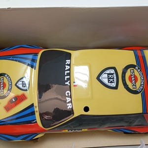May include: A yellow and black toy rally car with blue and red stripes. The car has the words "RALLY CAR" and "RADIOKONTROL ADVANCE" printed on it. The car is in a cardboard box.