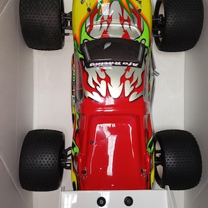 May include: A red, yellow, and silver remote-controlled car with large black tires. The car has a flame design on the hood and the text "Afa Racing". The car is in a white box with the text "READY-TO-RUN".