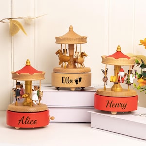May include: Three wooden carousel music boxes. Each has a red base and a wooden top. One carousel has the name "Alice" on the base, another has "Ella", and the third has "Henry". The carousel figures include horses, Santa, and other figurines.
