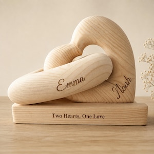 Op de afbeelding: Een lichtgekleurd houten sculptuur met twee in elkaar grijpende hartvormen op een rechthoekige voet. De naam Emma is gegraveerd op het voorste hart, Noah op de zijkant en de tekst Two Hearts, One Love staat op de voet.