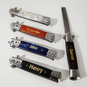 Könnte beinhalten: Eine Sammlung personalisierter Springkammkämme mit Namen wie "Leo", "Dylan", "Jake", "Henry" und "Gabriel" auf den Griffen. Die Kämme haben verschiedene Griffdesigns, darunter Weiß, Holzmaserung, Blau und Schwarz.