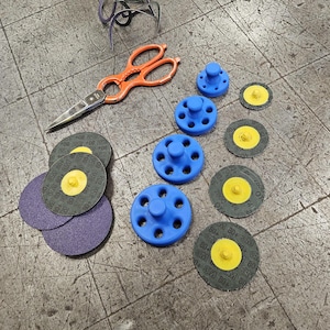 May include: Assortment of blue sanding discs with yellow centers and black sanding pads with yellow centers. The sanding discs and pads are arranged on a gray surface with a pair of orange scissors and a blue sanding disc holder.