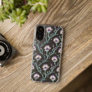 May include: A clear phone case with a floral design. The case features a repeating pattern of white and purple flowers with green stems on a black background.