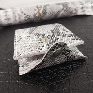 Może przedstawiać: Mały biały i czarny portfel ze skóry węża z zatrzaskiem.