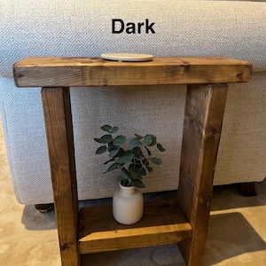 May include: A dark brown wooden side table with a shelf. The table has a rustic look and is made of reclaimed wood. The table is next to a white sofa with a grey patterned fabric. The table has a small plant in a white ceramic pot on the shelf.