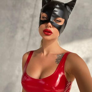 Puede incluir: Una máscara de gato de cuero negro con orejas puntiagudas. La persona lleva una camiseta sin mangas roja brillante y tiene pintalabios rojo. El fondo es una pared gris claro.