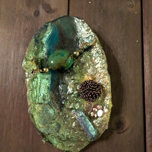 Op de afbeelding: Een handgemaakte, ovale decoratieve schaal met een getextureerd, metallic groen en goud oppervlak. Versierd met een grote groene steen, kleine gouden kralen, een donkerbruin bloemaccent en een blauwgroen kristal.