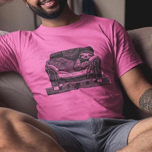 Puede incluir: Una camiseta rosa con una ilustración en blanco y negro de un perezoso en un sofá. La persona está sentada en un sofá beige con un cojín amarillo. La persona también sostiene un vaso de agua.