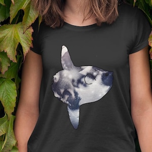 May include: Black t-shirt featuring a graphic of a sunfish, with a cloudy sky design. The shirt is worn by a person standing in front of a wall of green leaves. The person is wearing light blue jeans and bracelets.