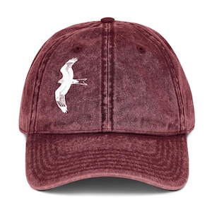 May include: A maroon baseball cap with a white graphic of a bird in flight. The cap has a curved brim and an adjustable strap. The hat is made of a soft, washed cotton material.