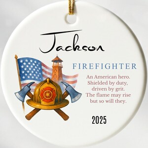 Op de afbeelding: Ronde witte keramische kerstversiering met een gouden ophangkoord. Het ontwerp toont een Amerikaanse vlag, een brandweerkazerne, een brandweerhelm en gekruiste bijlen. De tekst bevat "Jackson", "FIREFIGHTER" en het jaar "2025".
