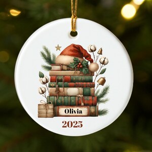 May include: Round white Christmas tree decoration featuring a festive design. A stack of books is topped with a Santa hat, holly, and cotton. The name "Olivia" and the year "2025" are printed on the ornament.
