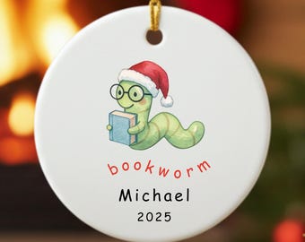 Puede incluir: Adorno navideño redondo y blanco con un gusano de libro de dibujos animados que lleva un gorro de Papá Noel y gafas, sosteniendo un libro. La palabra "bookworm" está escrita en rojo, con el nombre "Michael" y el año "2025" debajo.