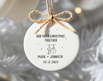 May include: A white ceramic Christmas ornament with the text "OUR FIRST CHRISTMAS TOGETHER", two hanging stockings, and the names "MARK + JENNIFER" with the date "25.12.2025". The ornament is suspended by a twine bow.