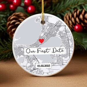 May include: Round ceramic ornament with a map design, a red heart, and the text "Our First Date" with the date 01.02.2025. The ornament hangs from a gold string, with a festive background of pine needles, berries, and pine cones.