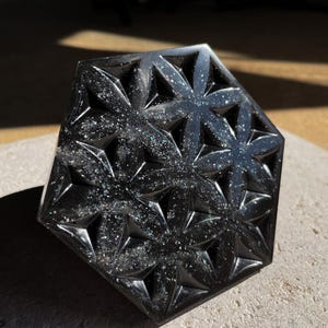 May include: A black, hexagonal decorative object with a floral pattern. The object has a glossy finish and is speckled with iridescent glitter. The design features repeating star-shaped flowers.