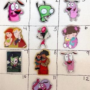 May include: A collection of twelve numbered acrylic pins featuring cartoon characters including Courage the Cowardly Dog, Gir from Invader Zim, Kim Possible, Ron Stoppable, Boo from Monsters Inc, and other animated figures with glittery finishes.