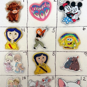 May include: A grid of twelve glittery acrylic character charms featuring a brown highland cow, a heart with the text I TRUST IN GOD, Mickey and Minnie Mouse, Coraline, Kim Possible, SpongeBob SquarePants, Precious Moments figures, and Pinky and the Brain.