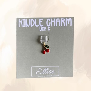 May include: A cherry charm with red enamel cherries and green leaves, attached to a clear USB-C connector. The charm is displayed on a grey card with the text "KINDLE CHARM USB C" and the brand name "Ellise Gifts & Design".