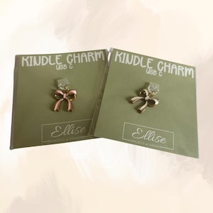 May include: Two Kindle charms with bow designs. One bow is pink and gold, the other is white and gold. Each charm is attached to a USB-C connector and displayed on a green card with the text "Kindle Charm USB C" and "Ellise Gifts & Design."