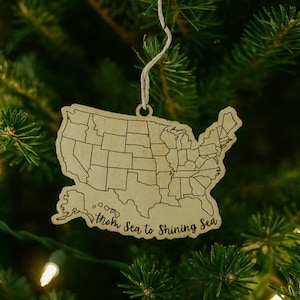 Puede incluir: Adorno navideño de madera con la forma de los Estados Unidos, con contornos de los estados y la frase "From Sea to Shining Sea". El adorno cuelga de una cuerda blanca sobre un fondo de agujas de pino verdes.