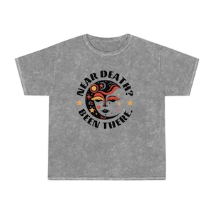 May include: A grey t-shirt with a distressed look featuring a black and white graphic of a crescent moon with a face and stars. The text "Near Death? Been There." is printed in black around the moon.