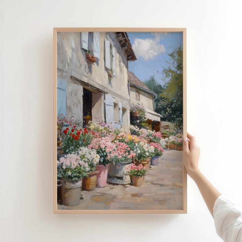 May include: A framed painting of a flower shop with a stone building, blue shutters, and a white awning. Potted flowers in pink, red, and white line the walkway. The artwork is in a light wooden frame.