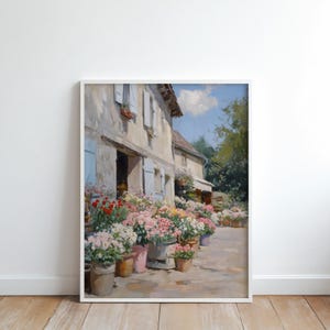 May include: A framed painting of a flower shop with a stone facade and blue shutters. Potted flowers in pink, red, and white are displayed outside. The artwork captures a sunny day in a European village, with a charming, idyllic scene.