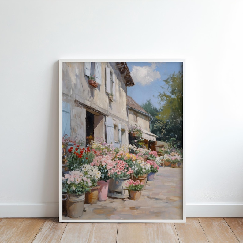 May include: A framed painting of a flower shop with a stone facade and blue shutters. Potted flowers in pink, red, and white are displayed outside. The artwork captures a sunny day in a European village, with a charming, idyllic scene.