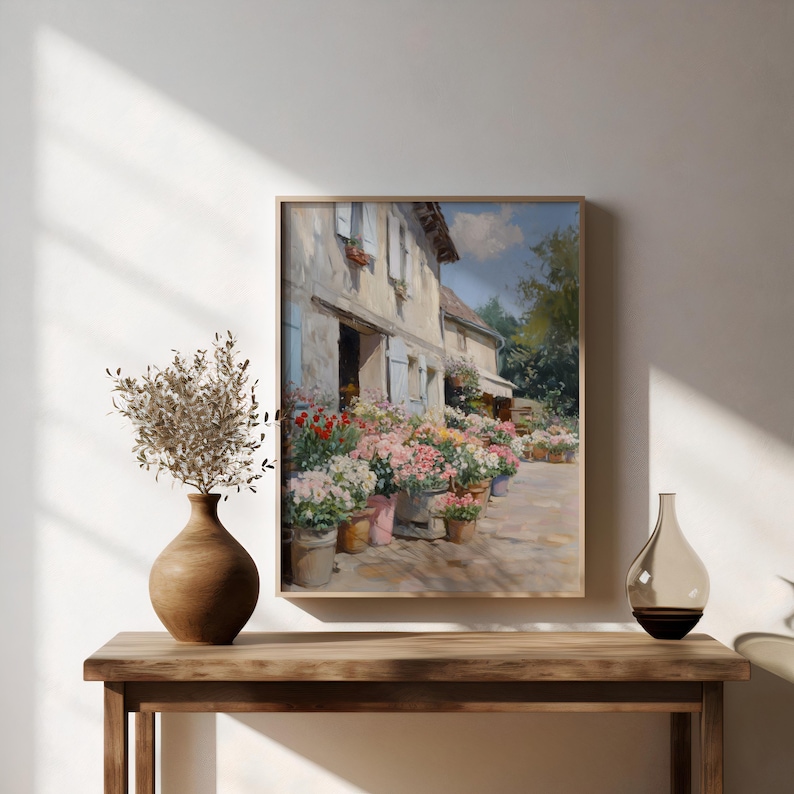 May include: A framed painting shows a charming flower shop with a stone facade, blue shutters, and flower pots. The artwork is displayed on a wooden table, with a brown vase of dried foliage and a decorative glass bottle.