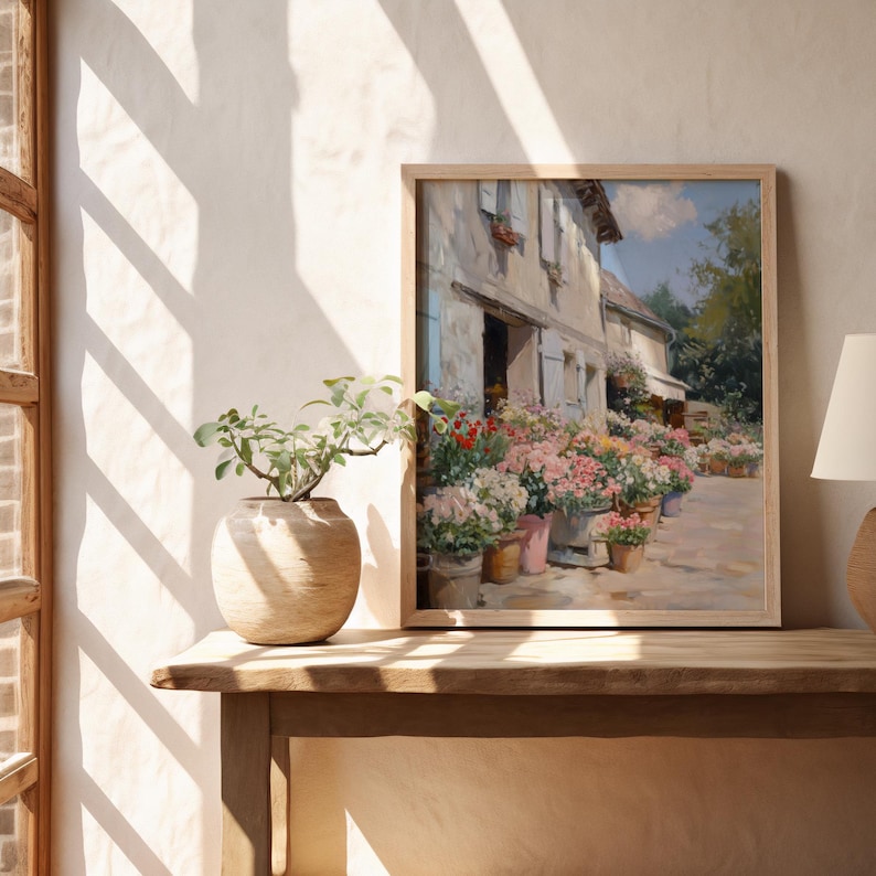 May include: A framed painting of a flower shop in a light wood frame. The artwork shows a building with white walls and blue shutters, with potted flowers in pink, red, and white. The painting is displayed on a wooden table.
