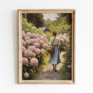 Op de afbeelding: Een ingelijste kunstdruk van een vrouw die loopt op een pad omzoomd met roze hortensia's. Ze draagt een witte blouse, een blauwe rok en een gevlochten tas. Het kunstwerk heeft een vintage stijl en een goudkleurige lijst.