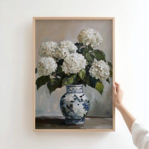 Puede incluir: Un jarr&oacute;n de porcelana blanca con dise&ntilde;os florales azules y verdes contiene un ramo de hortensias blancas. El jarr&oacute;n est&aacute; sobre una mesa de madera sobre un fondo gris claro.