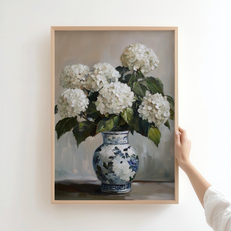 Puede incluir: Un jarr&oacute;n de porcelana blanca con dise&ntilde;os florales azules y verdes contiene un ramo de hortensias blancas. El jarr&oacute;n est&aacute; sobre una mesa de madera sobre un fondo gris claro.
