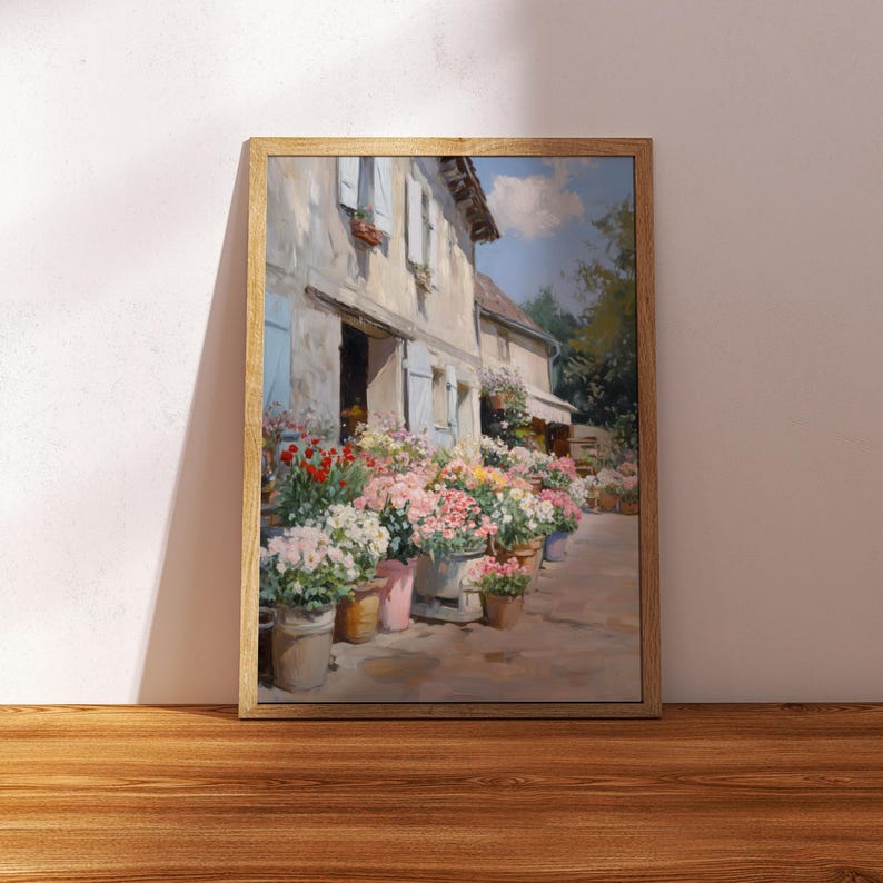 May include: A framed painting of a flower shop with a variety of colorful blooms in pots. The shop has light blue shutters and a weathered exterior. The artwork is set against a white wall and a wooden floor.
