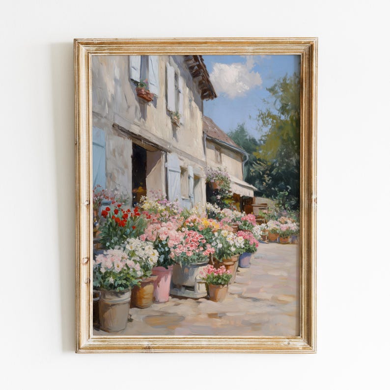 May include: A framed painting of a flower shop with a stone building, blue shutters, and numerous potted flowers in pink, red, and yellow. The artwork creates a tranquil, sunlit scene, with a view of a blue sky and a hint of greenery.