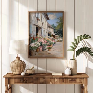 May include: A framed painting of a flower shop with vibrant flowers in pots. The artwork, in a wooden frame, is displayed on a wooden table with a lamp and decorative items. The scene is set against a white panelled wall.