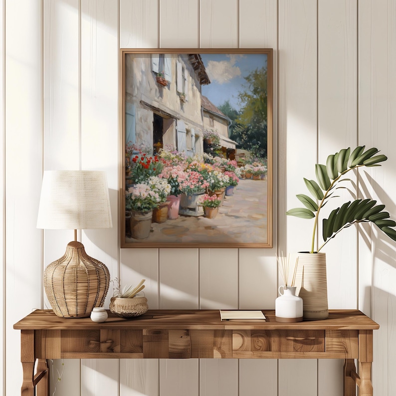 May include: A framed painting of a flower shop with vibrant flowers in pots. The artwork, in a wooden frame, is displayed on a wooden table with a lamp and decorative items. The scene is set against a white panelled wall.