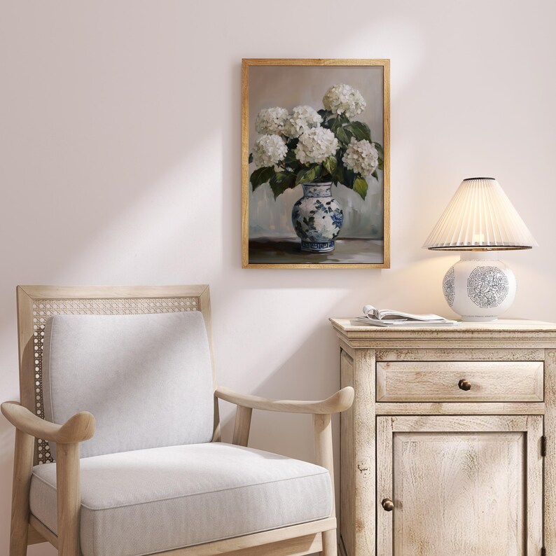 Puede incluir: Una pintura enmarcada de un ramo de hortensias blancas en un jarr&oacute;n de porcelana azul y blanco. El jarr&oacute;n est&aacute; sobre una mesa con una superficie marr&oacute;n claro. La pintura est&aacute; colgada en una pared blanca.
