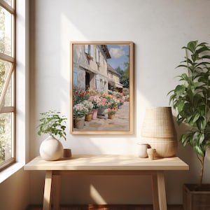 May include: A framed painting of a flower shop with vibrant flowers in pots along the pavement. The building features light blue shutters and a pale exterior. The artwork is displayed on a light wood table, alongside a white vase and a woven basket.