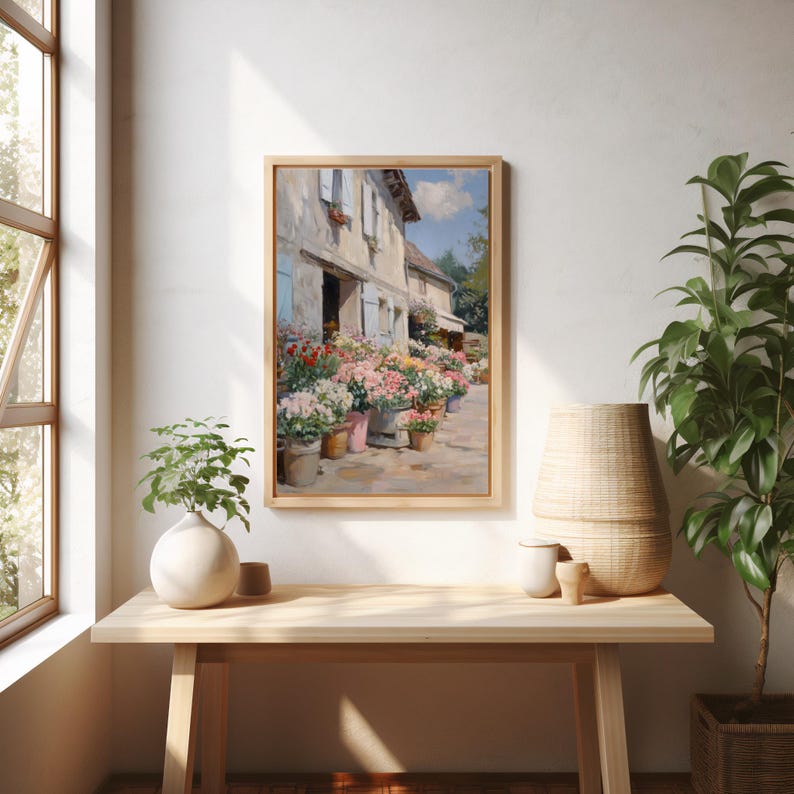 May include: A framed painting of a flower shop with vibrant flowers in pots along the pavement. The building features light blue shutters and a pale exterior. The artwork is displayed on a light wood table, alongside a white vase and a woven basket.