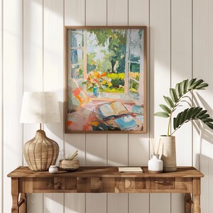 May include: A painting depicting a window with a view of a garden. The window is open and there are books on a table in front of it. The painting is in a wooden frame and is hanging on a white wall.
