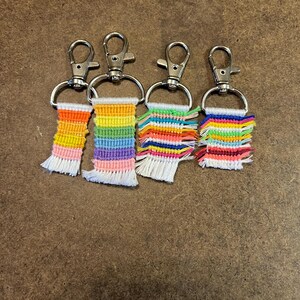 May include: Four handmade keychains with rainbow-colored woven designs. Each keychain features a silver-colored clasp and a rectangular woven section with a fringe at the bottom. The colors include orange, yellow, green, blue, and purple.