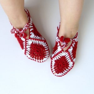 Può includere: Ciabatte in crochet rosso e bianco con un design decorativo. Le ciabatte hanno un dettaglio a pompon sulla parte superiore.